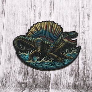 May include: A black and blue embroidered patch of a Spinosaurus dinosaur with a large sail on its back, standing in water with waves.