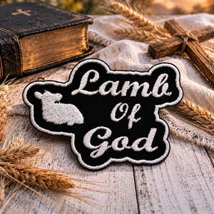 May include: Black embroidered patch with the words "Lamb Of God" and a white lamb silhouette. The patch is surrounded by a white border. A wooden cross, a Bible, and wheat stalks are in the background.