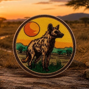 May include: A circular embroidered patch featuring a spotted African wild dog standing in a grassy savanna landscape. The design includes a bright orange sun set against a yellow sky, with green bushes and a dark brown stitched border edge.