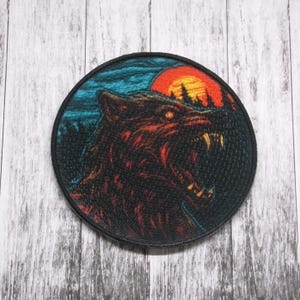 Może przedstawiać: Okrągła naszywka haftowana z czarnym wilkiem wyjącym do czerwono-pomarańczowego zachodu słońca. Oczy wilka świecą na czerwono, a tło jest ciemnoniebieskie z kilkoma białymi chmurami.