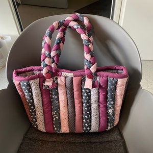 Peut inclure: Un sac cabas matelass&eacute; avec une anse en tissu tress&eacute;. Le sac est fait de diff&eacute;rents tons de rose et de violet avec des motifs floraux. Le sac est pos&eacute; sur une chaise grise.