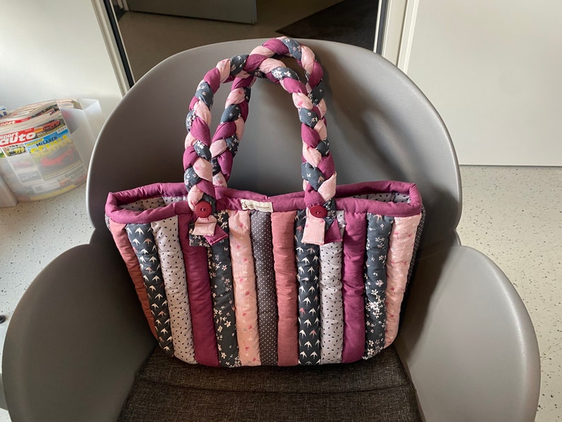 Peut inclure: Un sac cabas matelass&eacute; avec une anse en tissu tress&eacute;. Le sac est fait de diff&eacute;rents tons de rose et de violet avec des motifs floraux. Le sac est pos&eacute; sur une chaise grise.