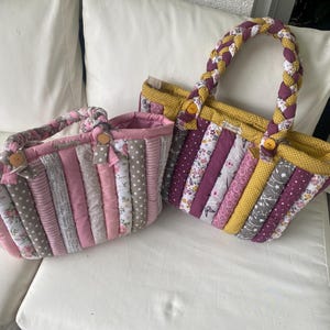 Könnte beinhalten: Zwei gesteppte Tragetaschen mit geflochtenen Griffen. Eine Tasche ist rosa und grau mit Blumen- und Tupfenmustern. Die andere ist lila, gelb und grau mit Blumen- und Sternmustern. Beide Taschen haben Holzknopfverschlüsse.