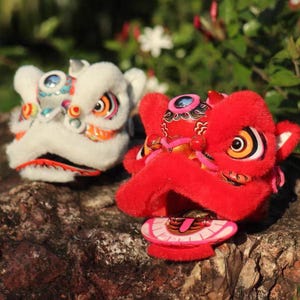 Puede incluir: Dos cabezas de león de peluche, una blanca y otra roja, descansan sobre una superficie rocosa texturizada. El león blanco tiene detalles en naranja, azul y negro. El león rojo presenta detalles en rosa y dorado, con ojos grandes y expresivos.