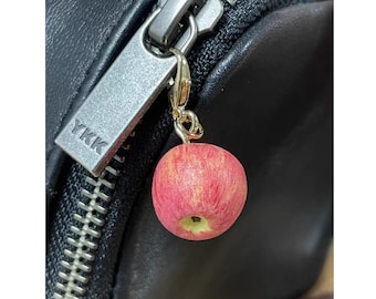 May include: A black leather bag with a silver zipper pull. The zipper pull has a small, red apple charm attached to it. The charm is made of a soft, textured material and has a gold-coloured stem.