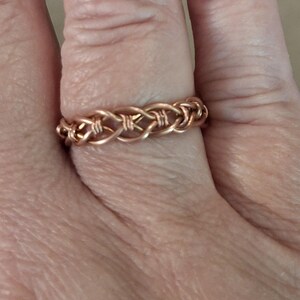 May include: A copper-colored ring featuring a woven wire design. The band is constructed from multiple strands of metal wire intertwined to create a continuous, repeating pattern that encircles the finger with a consistent, textured appearance.