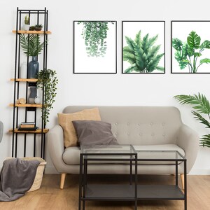 K&ouml;nnte beinhalten: Ein Wohnzimmer-Interieur mit einem grauen Sofa, einem schwarzen Couchtisch aus Metall, einem grauen Sessel, einem schwarzen Regal aus Metall mit Holzb&ouml;den und drei gerahmten Drucken von tropischen Pflanzen an einer wei&szlig;en Wand.