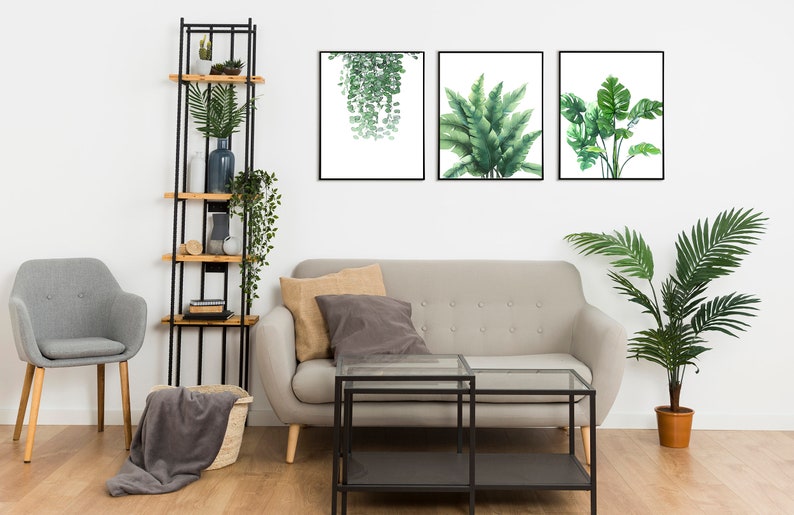 K&ouml;nnte beinhalten: Ein Wohnzimmer-Interieur mit einem grauen Sofa, einem schwarzen Couchtisch aus Metall, einem grauen Sessel, einem schwarzen Regal aus Metall mit Holzb&ouml;den und drei gerahmten Drucken von tropischen Pflanzen an einer wei&szlig;en Wand.