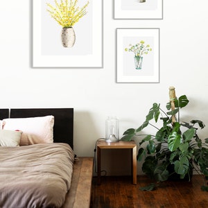 May include: Three framed watercolor paintings of yellow flowers in different vases. The paintings are hanging on a white wall above a wooden bed frame and a small wooden side table with a lamp.