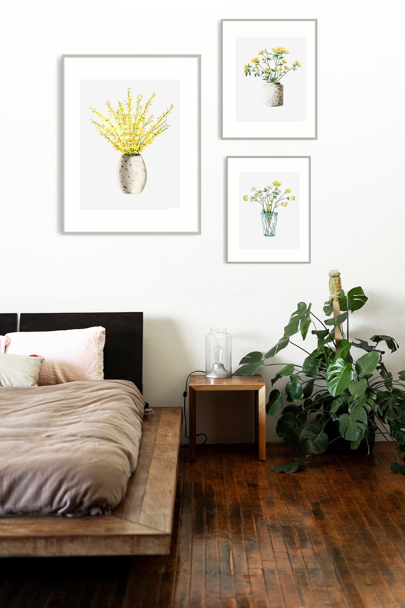 May include: Three framed watercolor paintings of yellow flowers in different vases. The paintings are hanging on a white wall above a wooden bed frame and a small wooden side table with a lamp.