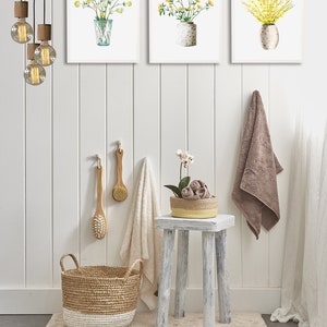 May include: A bathroom interior with a white, wood-paneled wall, a white stool, a woven basket, and three framed watercolor paintings of yellow flowers in vases.