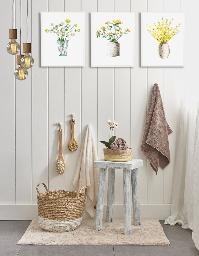 May include: A bathroom interior with a white, wood-paneled wall, a white stool, a woven basket, and three framed watercolor paintings of yellow flowers in vases.