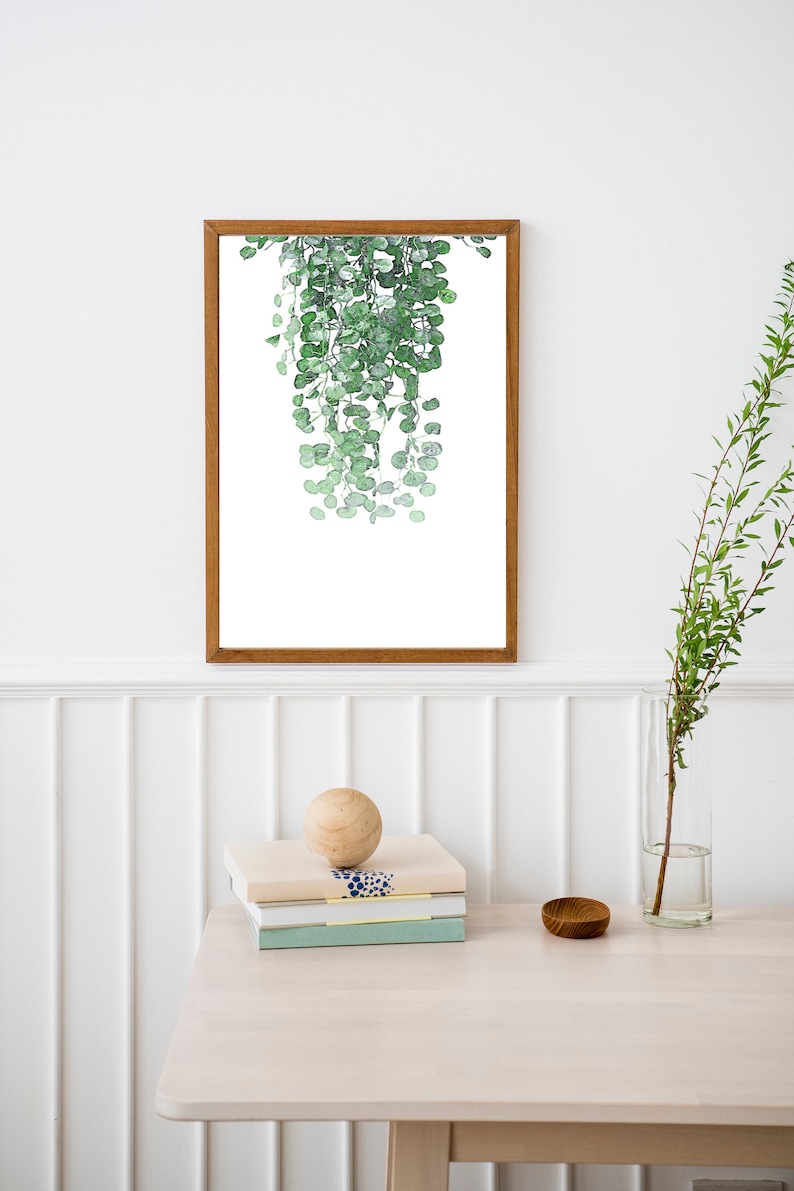 K&ouml;nnte beinhalten: Ein gerahmter Aquarelldruck einer h&auml;ngenden Pflanze mit gr&uuml;nen Bl&auml;ttern vor wei&szlig;em Hintergrund. Der Druck ist an einer wei&szlig;en Wand &uuml;ber einem hellen Holztisch mit einer Holzkugel, einem Stapel B&uuml;chern und einer kleinen Holzschale angebracht.