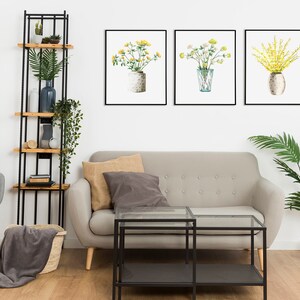 May include: A living room interior with a gray sofa, a gray armchair, a black metal shelf with wooden shelves, a glass coffee table, a potted palm tree, and three framed watercolor paintings of yellow flowers in vases.