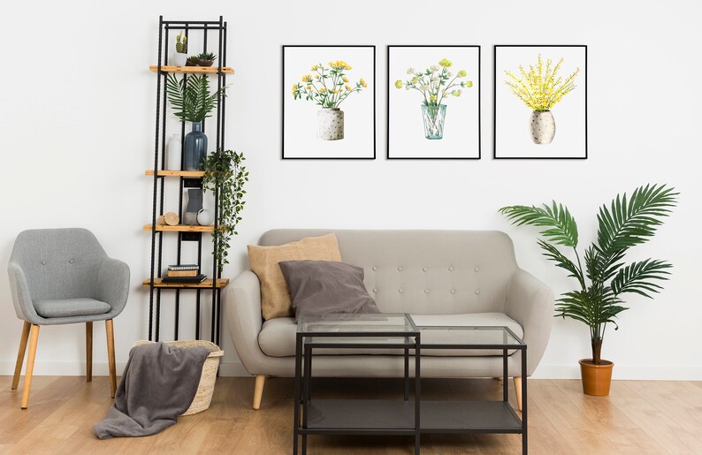 May include: A living room interior with a gray sofa, a gray armchair, a black metal shelf with wooden shelves, a glass coffee table, a potted palm tree, and three framed watercolor paintings of yellow flowers in vases.