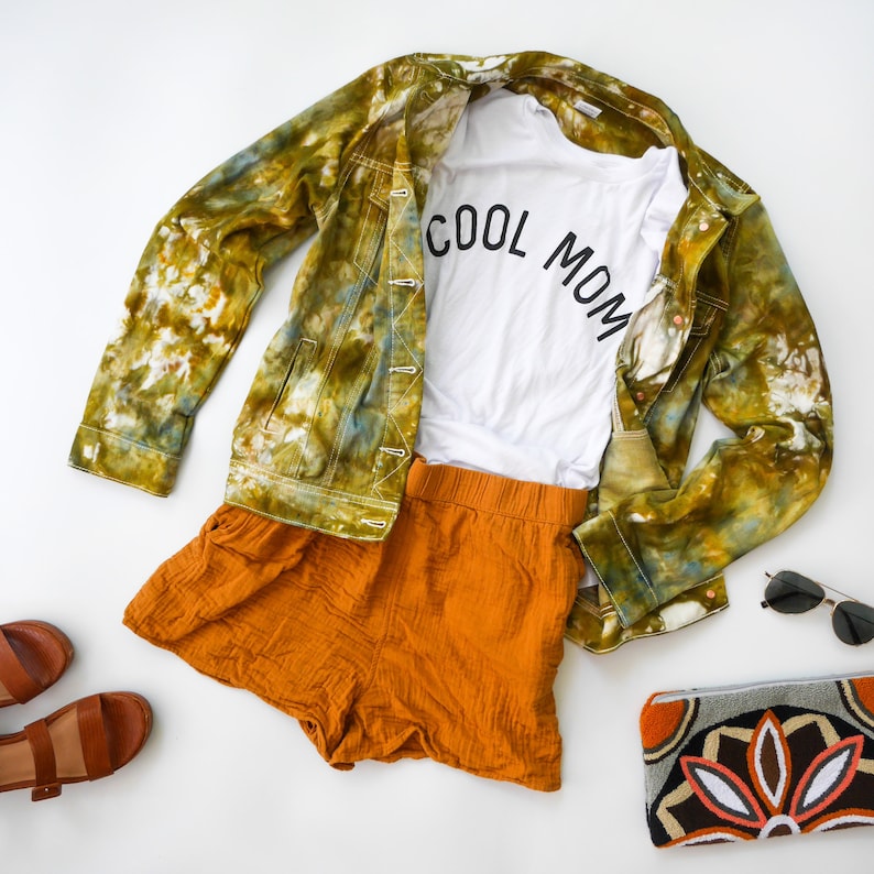 May include: A green and brown tie-dye denim jacket, a white t-shirt with the text "COOL MOM" in black, orange shorts, and a gray and brown clutch with a floral design.