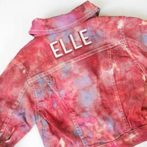 May include: A red, pink, and purple tie-dye denim jacket with white stitching. The back of the jacket has the name "ELLE" embroidered in white.