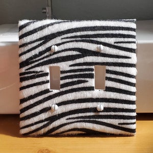 Peut inclure: Un cache-interrupteur double décoratif avec un motif zébré. Le couvercle présente un fond blanc avec des rayures noires, deux ouvertures rectangulaires pour les interrupteurs et quatre trous de vis. Les vis sont blanches.