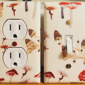 Puede incluir: Dos cubiertas de enchufes y una cubierta de interruptor de luz con un fondo crema y un patrón repetido de setas en tonos rojos, marrones y morados. Los enchufes y el interruptor son blancos.