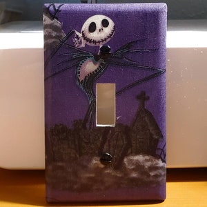 Peut inclure: Un cache-interrupteur violet avec une image en noir et blanc de Jack Skellington du film L'Étrange Noël de monsieur Jack. L'image montre Jack Skellington avec son sourire caractéristique et ses longs membres fins.