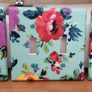 May include: Three light switch covers with a floral pattern. The covers are turquoise with a white background and feature a variety of colorful flowers.