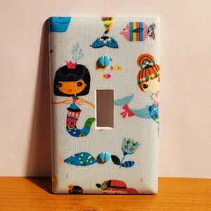 May include: A light switch cover with a blue and white fabric design featuring mermaids, fish, and flowers. The cover has two blue screws.