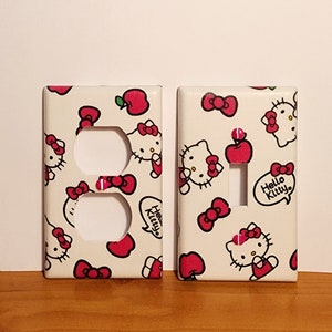 May include: Two white light switch and outlet covers with a Hello Kitty pattern. The pattern features Hello Kitty, red bows, and red apples on a white background. The text "Hello Kitty" is in a speech bubble on the light switch cover.