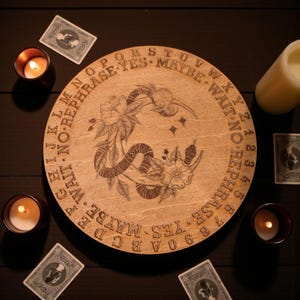 May include: A round wooden Ouija board with engraved letters, numbers, and phrases. The board features a floral and snake design in the center. The image includes lit candles and tarot cards, creating a mystical atmosphere.