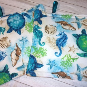 May include: Two square pot holders with a sea life pattern. The design features blue and green sea turtles, seahorses, seashells, starfish, and coral on a white and light blue background. Each pot holder has a teal loop.