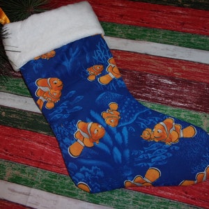 May include: A blue Christmas stocking featuring a pattern of orange and white clownfish swimming among blue coral and seaweed. The top of the stocking has a white, fluffy trim and a gold hanging loop.