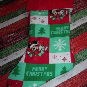 May include: Christmas stocking with a red plush cuff. The body features a patchwork design with Mickey Mouse, snowflakes, Christmas trees, and the words "MERRY CHRISTMAS" in green and red blocks. The background is a diagonal pattern of red, white, and green.