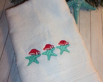 May include: White bath towel with three embroidered starfish wearing Santa hats. The starfish are turquoise and the hats are red.