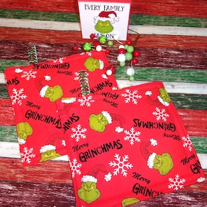 May include: Red Grinch-themed Christmas potholders with white snowflakes and the text "Merry Grinchmas." A small sign with the Grinch and the words "Every Family" is also visible. Decorative Christmas ornaments are in the background.