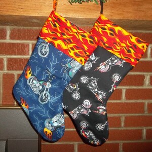 May include: Two Christmas stockings with motorcycle designs. One stocking is blue with motorcycle and flame graphics, the other is black with motorcycle graphics. Both have a red and yellow flame pattern at the top.