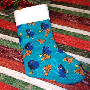 May include: A Christmas stocking with a teal background featuring cartoon images of blue and orange fish. The top of the stocking is white and fluffy. The stocking is on a striped background of red, white, and green.