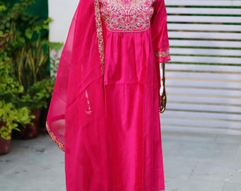 May include: A pink silk kurta with gold embroidery and a matching pair of pants. The kurta has a V-neckline and a gathered waist. The pants are straight-legged and have a gold embroidered trim. The outfit is displayed on a mannequin.