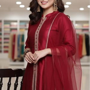 May include: A burgundy-colored formal dress with gold embroidery along the front and neckline. The dress has long sleeves and is paired with a sheer scarf. The woman is wearing gold earrings.