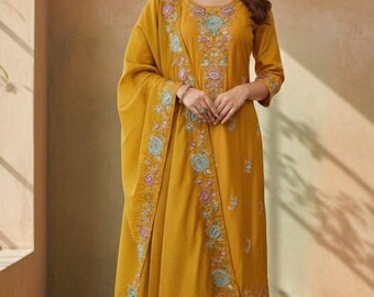Puede incluir: Conjunto salwar kameez bordado amarillo mostaza. El conjunto incluye una túnica, un pantalón y una bufanda a juego. La túnica y la bufanda presentan bordados florales en tonos azules, rosas y verdes. El conjunto se lleva con sandalias doradas.