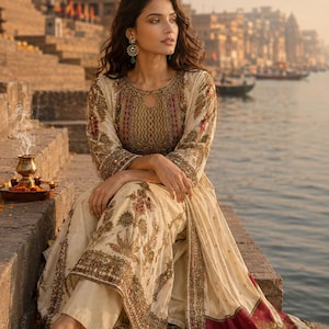 May include: A cream-colored long-sleeved tunic and matching trousers featuring intricate gold, red, and green floral embroidery. The tunic has a round neckline with a small keyhole cutout and a wide, decorative border along the hem.