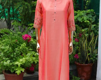 May include: A coral pink long-sleeved dress with floral embroidery on the cuffs. The dress has a round neckline and a button closure at the neck.