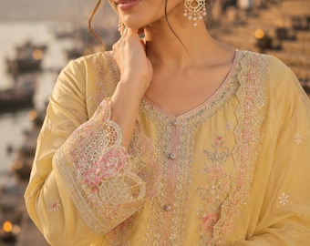 May include: A pale yellow long-sleeved tunic featuring intricate pink and white floral embroidery, a V-neckline with small buttons, and scalloped lace cuffs. The garment is paired with matching gold-toned chandelier earrings with pink beads.