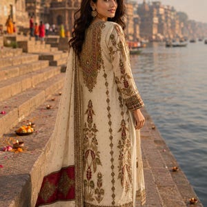May include: An off-white long-sleeved tunic and matching trousers featuring intricate gold, red, and green embroidery. A coordinating shawl with a deep red border and gold embellishments drapes over the shoulder, completing the ensemble.
