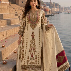 May include: A cream-colored long-sleeved tunic with intricate embroidery in shades of red, gold, and green, paired with matching trousers and a coordinating shawl featuring a wide maroon border with gold embellishments and tassel accents.