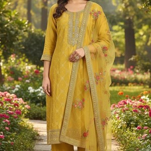 May include: Mustard yellow Indian salwar kameez suit. The outfit includes a long-sleeved tunic, matching pants, and a sheer dupatta scarf. The tunic features intricate embroidery around the neckline and cuffs. The dupatta is adorned with floral embroidery.