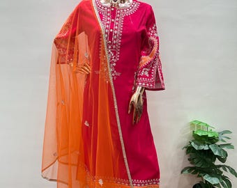 May include: A vibrant fuchsia-colored salwar kameez ensemble. The outfit features intricate white embroidery and orange accents on the sleeves and hem. An orange sheer dupatta with silver trim drapes over the outfit, creating a striking contrast. The outfit is displayed on a mannequin.