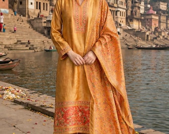 May include: A woman wearing a mustard-colored salwar kameez with a matching dupatta. The outfit features floral embroidery and a red and orange floral border. The background shows a river and buildings.