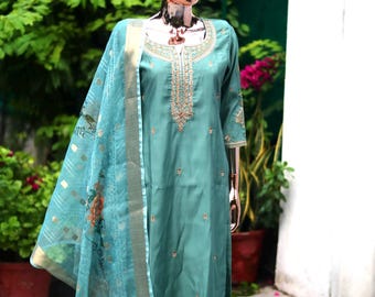 May include: A teal blue embroidered kurta with a matching pair of pants and a sheer dupatta with floral embroidery. The outfit is displayed on a mannequin against a backdrop of greenery.