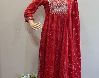 May include: A red Indian salwar kameez ensemble. The outfit includes a long-sleeved tunic with intricate embroidery around the neckline, matching pants, and a flowing scarf. The fabric features a subtle, repeating pattern.