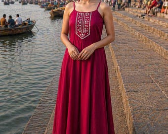 May include: A woman wearing a burgundy maxi dress with a detailed embroidered bodice. The dress has thin straps and a flowing skirt. She is standing on a stone walkway near a river with boats and buildings in the background.