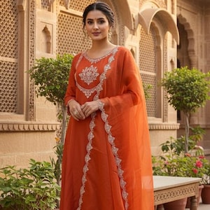May include: An orange-red Indian salwar kameez ensemble. The outfit includes a long-sleeved tunic with intricate white embroidery around the neckline and down the front, matching pants, and a sheer dupatta. The woman is standing in front of a sandstone building.